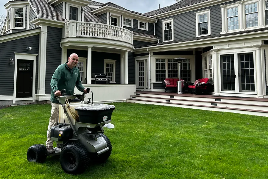 Lawn care professional operating a ride-on spreader across a vibrant green lawn in front of a large, elegant gray house.